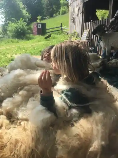 Zwei Kinder sitzen lachend in einem grossen Haufen frisch geschorener Wolle, hinten eine Ziege und die Wiese.
