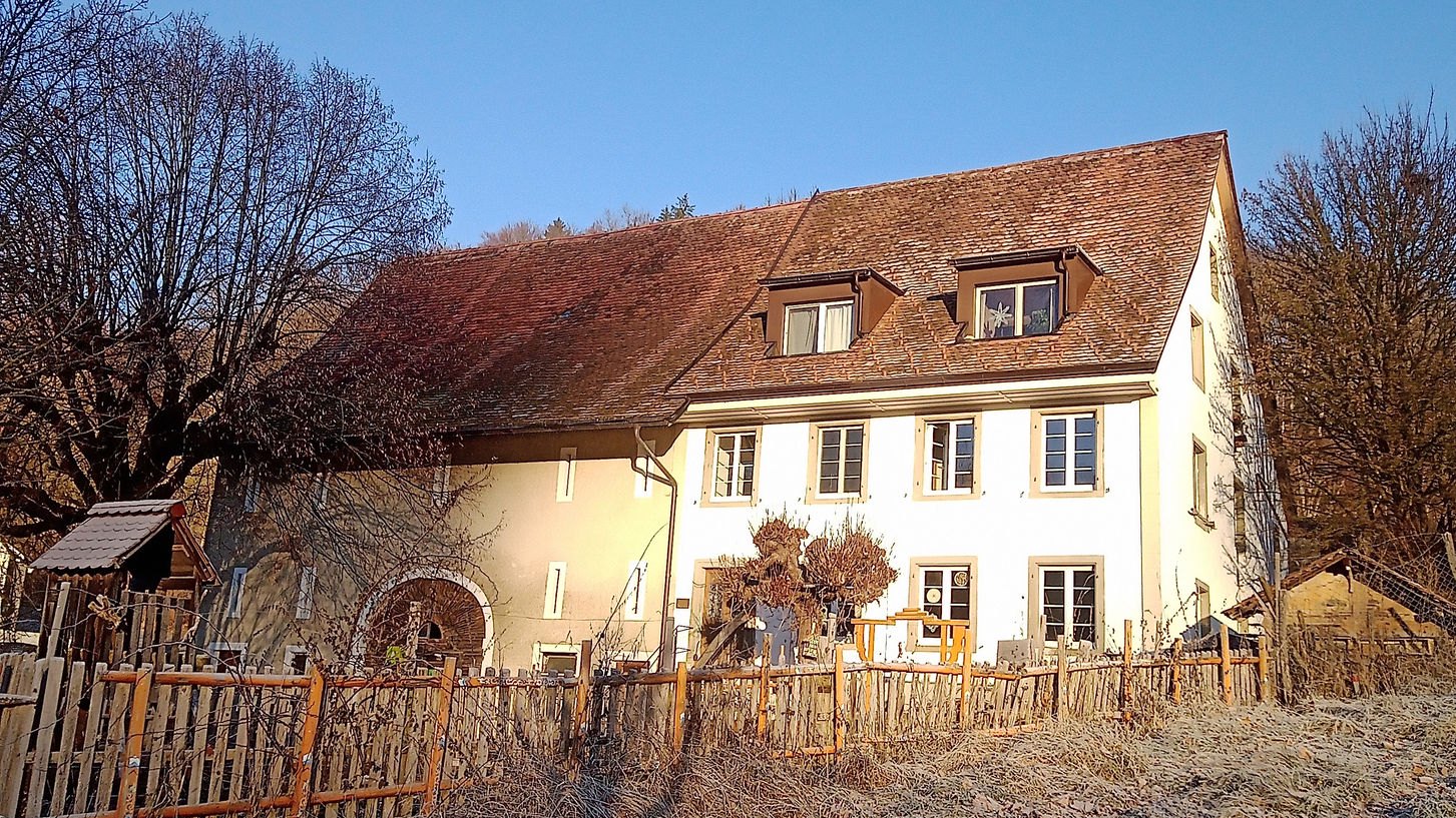 Das Hofhaus des Seehofs an einem klaren Wintermorgen. Ein langgestrecktes Bauernhaus mit weiss verputzter Fassade, Sprossenfenstern mit Läden und zwei Dachgauben, links geht es in den älteren Ökonomieteil mit einem gemauerten Rundbogen über. Davor ein Staketenzaun aus Haselruten und raureifbedecktes Gras.